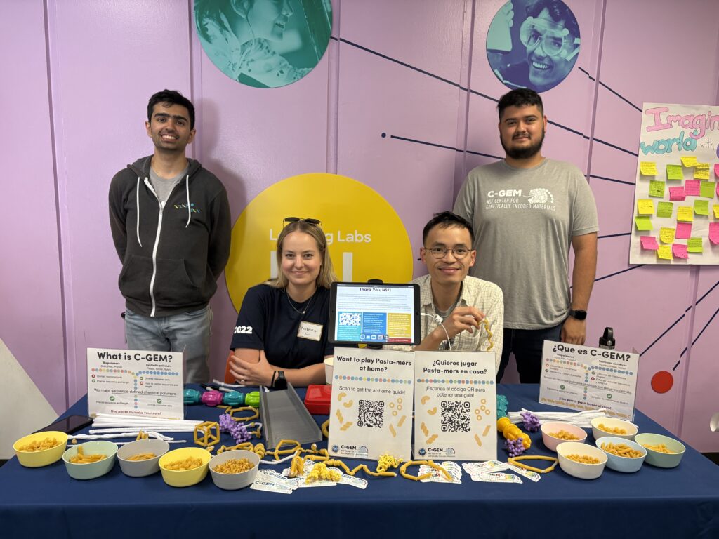 Four C-GEM scientists posing for a picture, sitting and standing behind a table. On the table are bowls of dried pasta, pipe cleaners, flyers that explain C-GEM science with QR codes, and examples of pasta crafts scattered across the table.