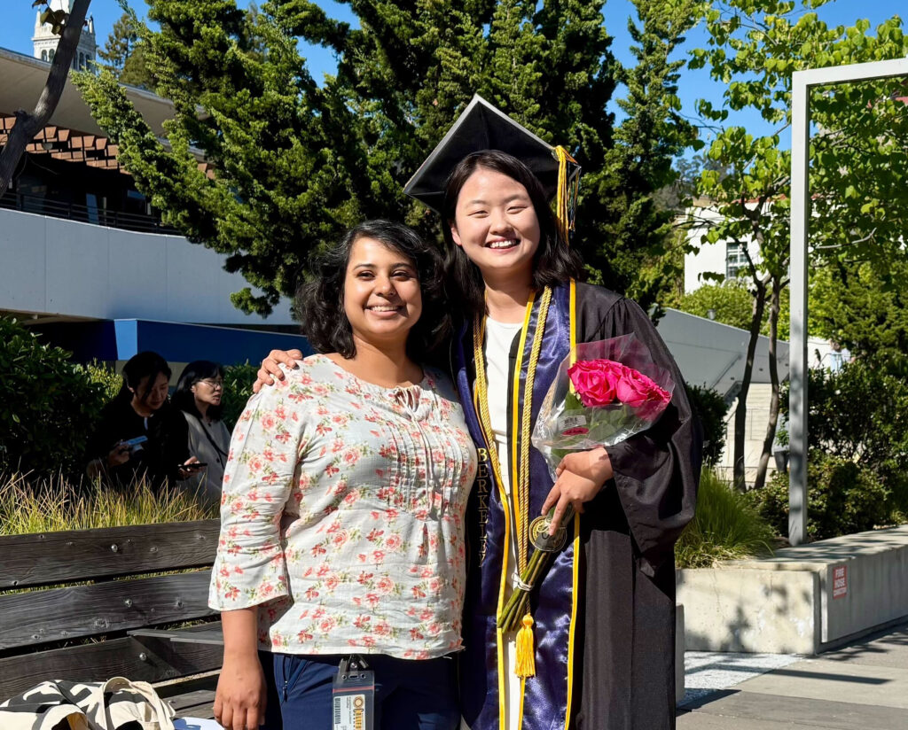 Cathy is wearing a black cap and gown with a blue and gold stole and gold cord around her neck. She is holding pink flowers. Chandrima is standing next to Cathy in a floral shirt. They are smiling at the camera and are in front of a building and trees.