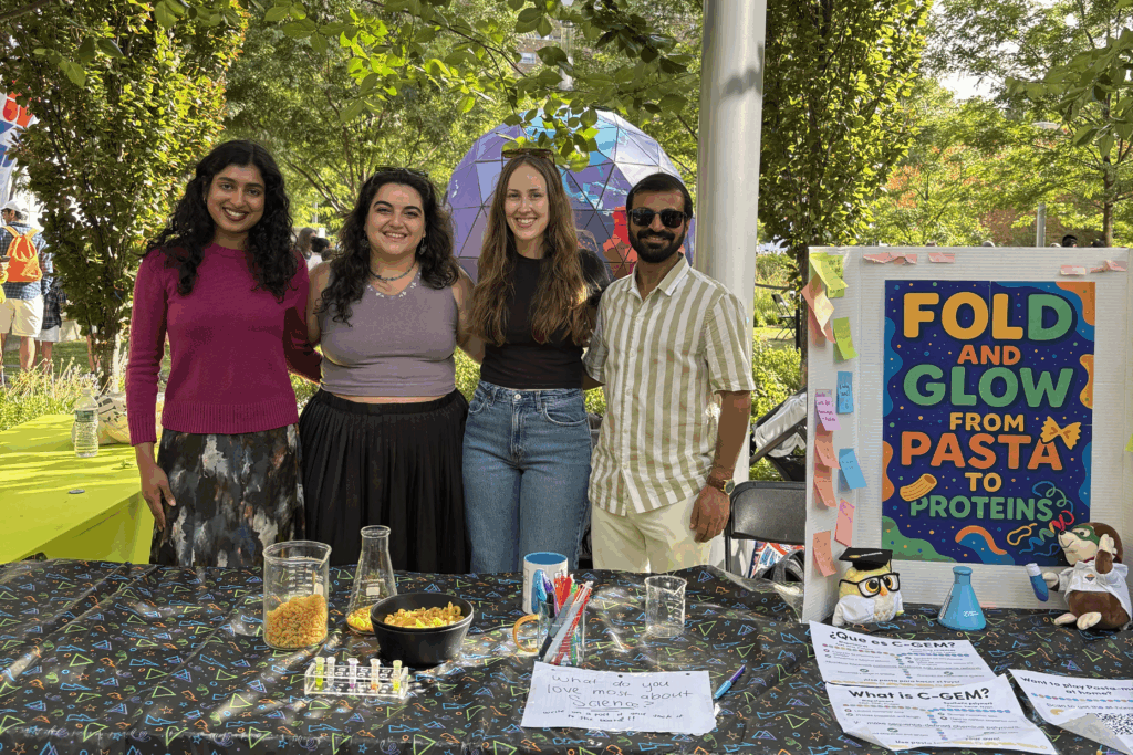 Four people smiling and standing behind a table covered in a black table cloth with a colorful shape pattern. On the table are beakers, flasks, and bowls filled with dried pasta, pens, a small rack with tubes containing colorful substances, some flyers that say "What do you love most about science" and "What is C-GEM?", and an upright trifold poster covered in post-it notes and a large colorful poster that says "Fold and Glow from Pasta to Proteins."