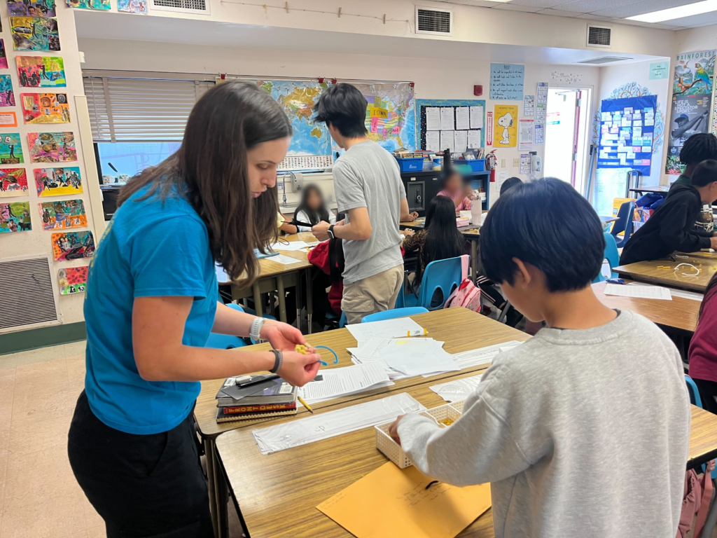A woman and an elementary schooler are leaning over a classroom desk, holding dry pasta and pipe cleaners.