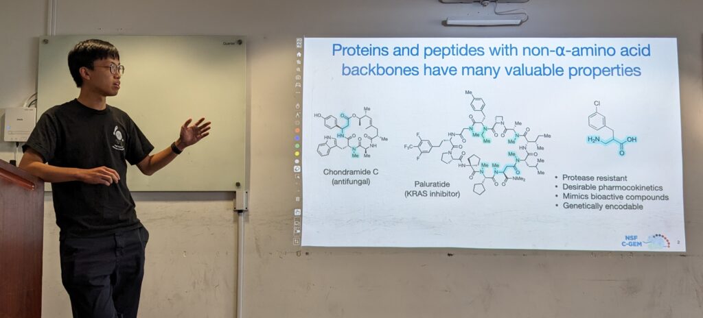 Thomas stands,mid-gesture, to the left of the presentation. The slide title reads "Proteins and peptides with non-alpha-amino acid backbones have many valuable properties" with bullets underneath reading: protease resistant, desirable pharmocokinetics, mimics bioactive compounds, genetically encodable.
